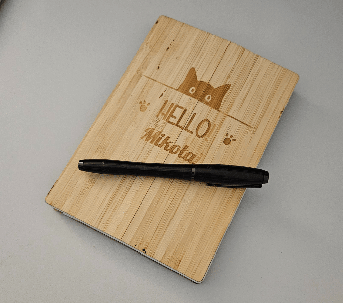 dziennik z drewnianą okładką i rysunkiem kota z napisem: "HELLO Mikołaj", po obu stronach napisu są łapki kota. Na dzienniku leży czarne pióro.